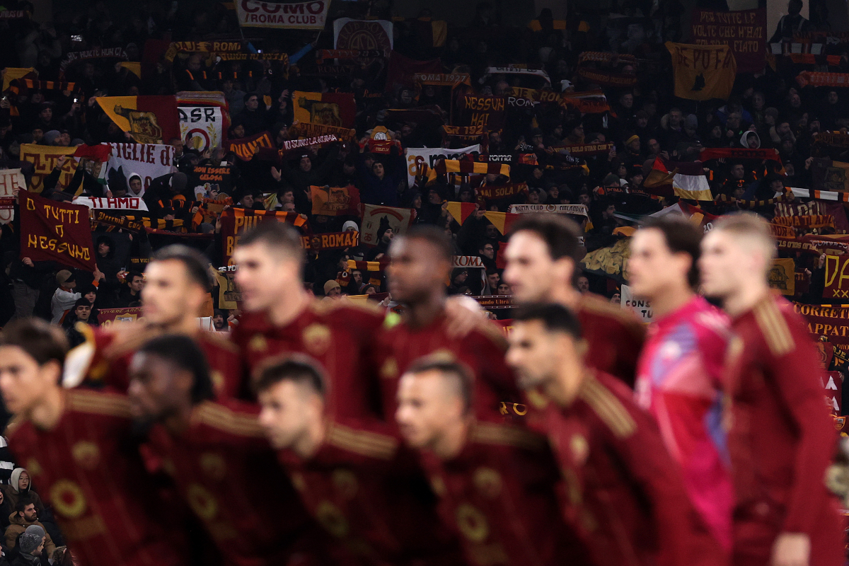 Roma players line up prior to their match against Tottenham Hotspur with a view of their fans in the background.