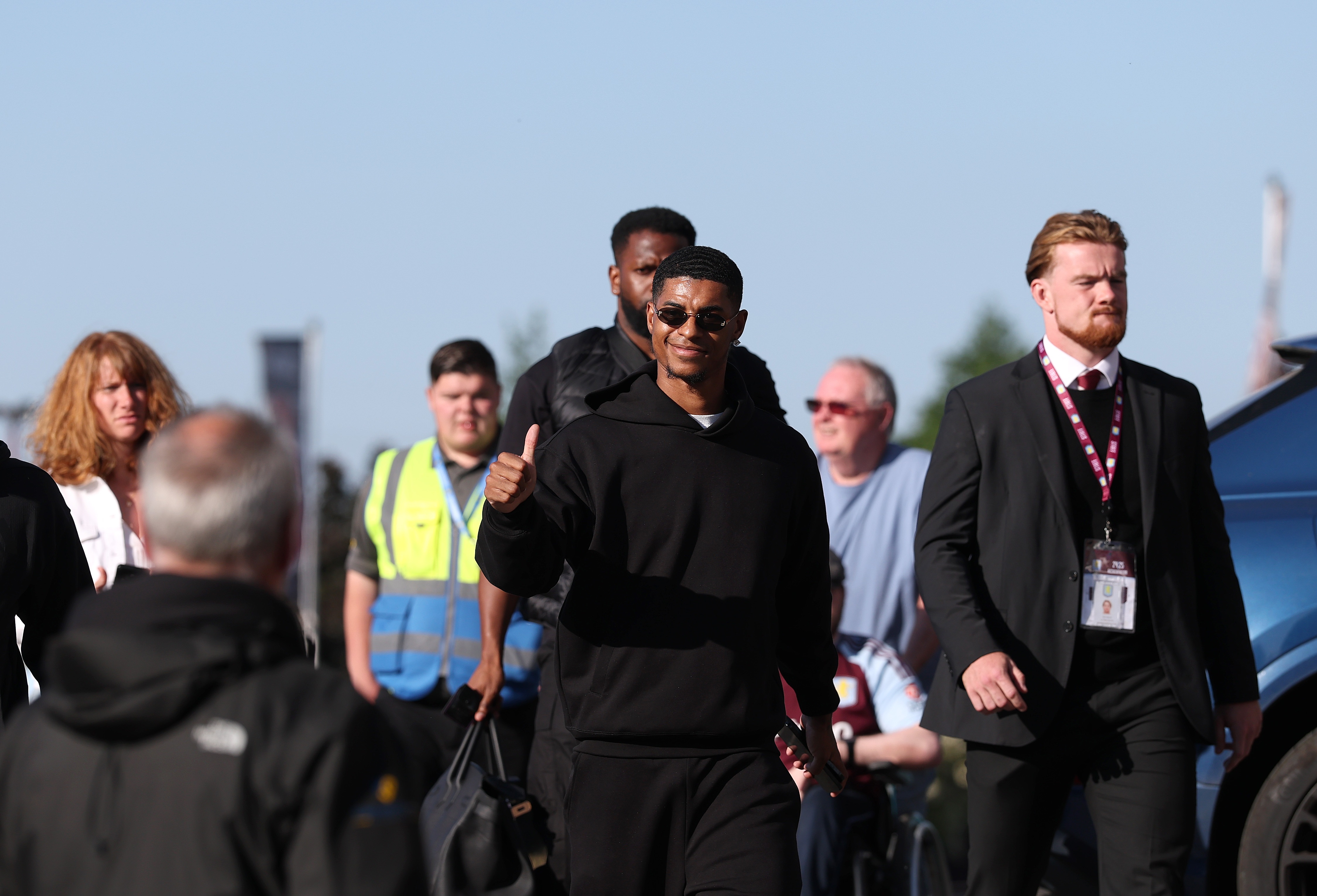 Marcus Rashford gives a thumbs-up whilst walking with a security detail.